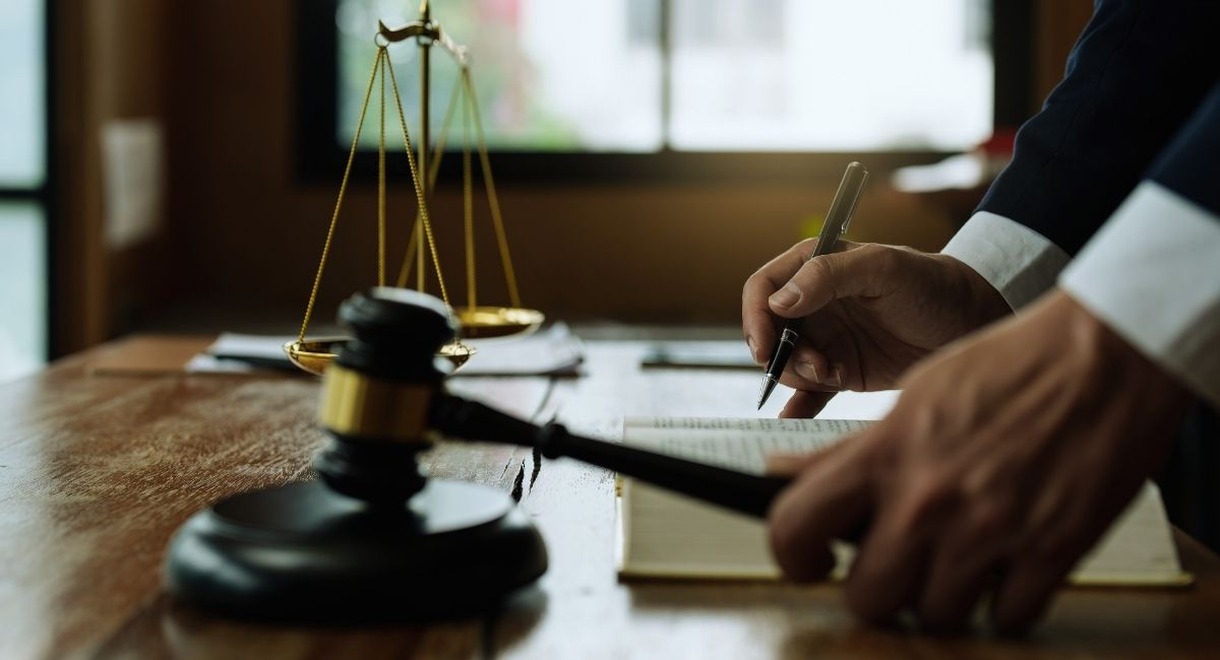 Main d'un avocat annotant un ouvrage de droit, marteau de justice et balance poses sur le bureau