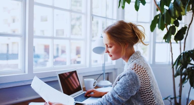 Femme lisant un document papier devant son ordinateur portable, près d'une fenêtre lumineuse